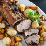A pork loin roll sits sliced on a white platter with roasted potatoes and carrots around it.