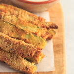 Keto air fryer zucchini fries sit on a cutting board next to a small bowl of dip.