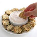 Air Fried Pickles on a white platter with a bowl of dip in the center. A hand dips one pickle into the dip.