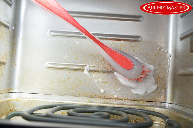 A red brush scrubs the inside of an air fryer with a baking soda solution.
